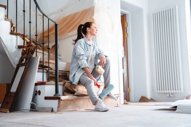 A young woman joyfully sits against the background her home remodeling.