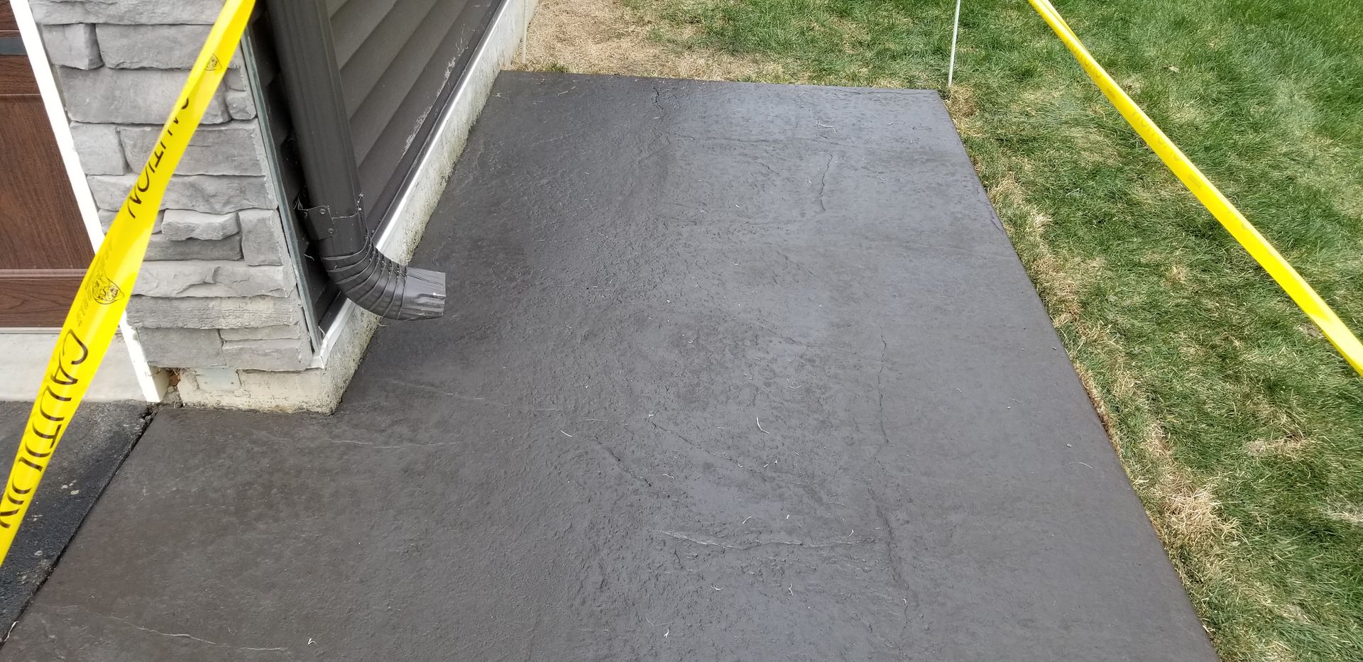 Freshly poured dark concrete sidewalk next to a house with a gutter and caution tape.