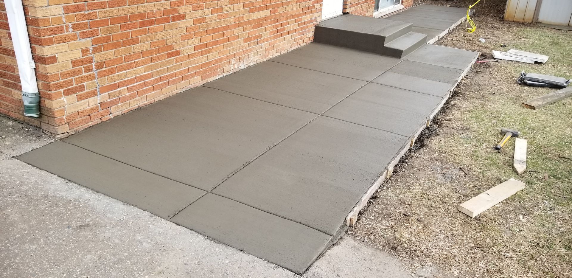 Newly poured concrete sidewalk and steps next to a brick wall. Construction tools are nearby.