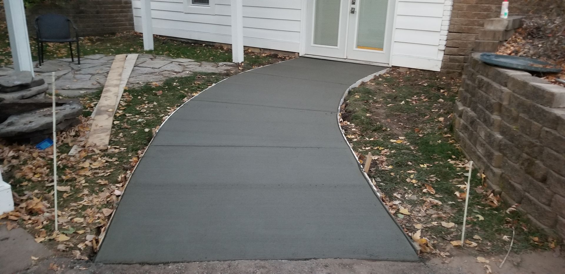 Newly poured concrete walkway curves towards a white building with double doors; autumn leaves scattered around.