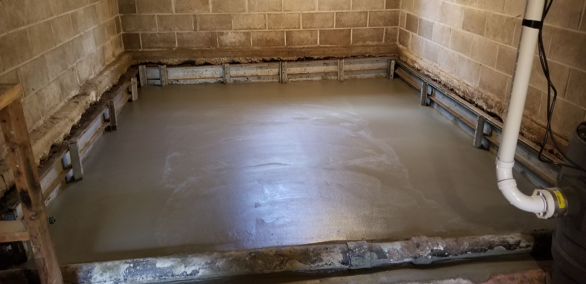Freshly poured concrete floor inside a room with brick walls and metal framework. A white pipe is on the right side.