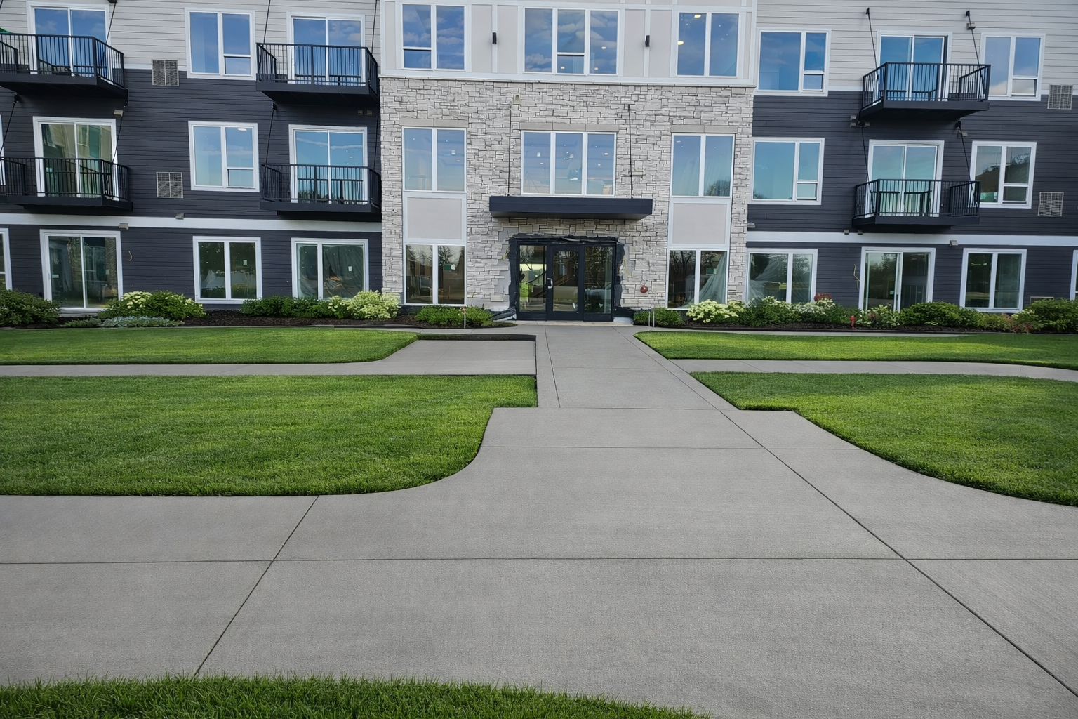 Gray concrete walkway leading to an apartment building entrance, flanked by green grass.