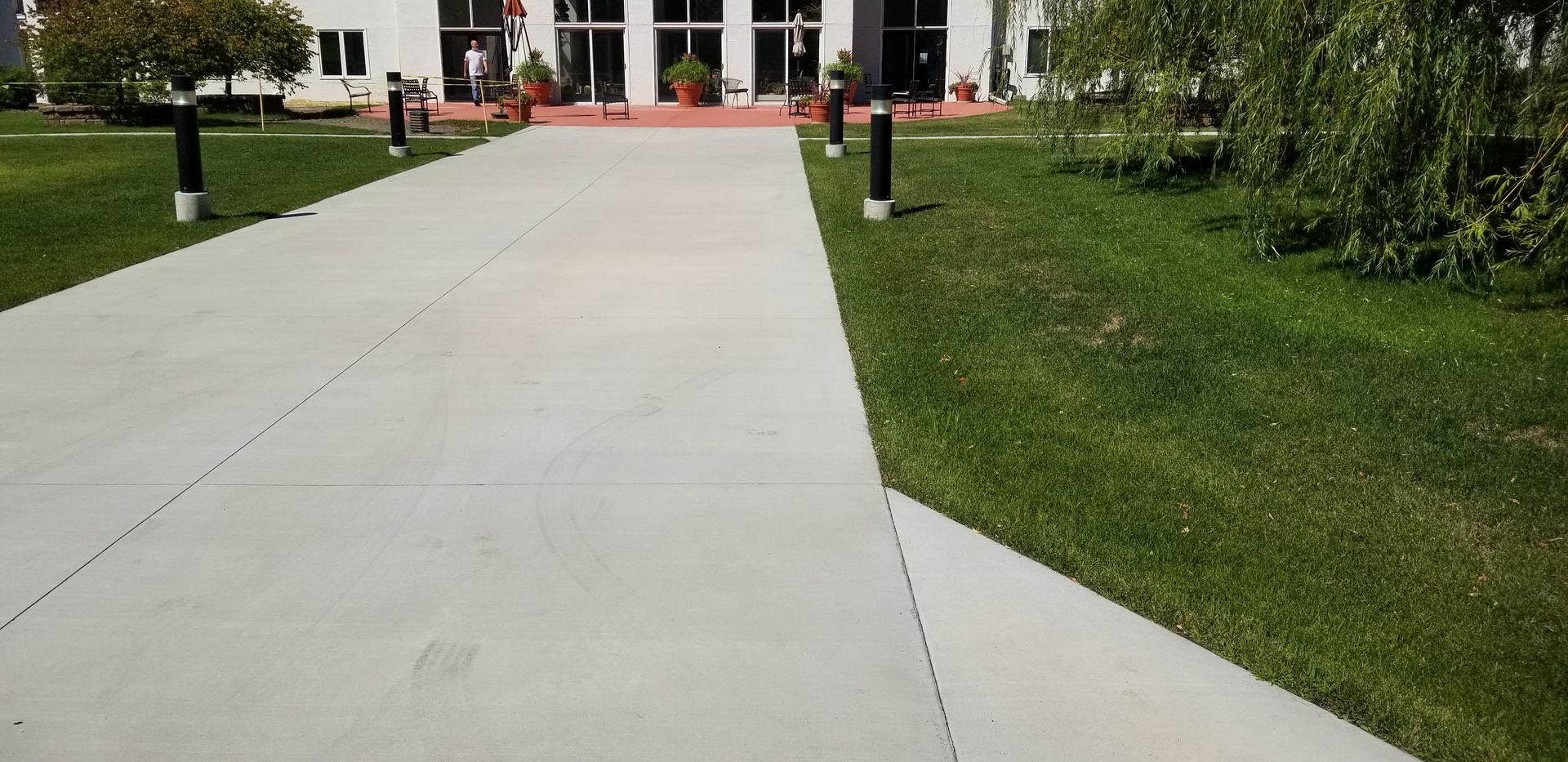 Concrete pathway leading to a building with glass doors and a red trim, flanked by grass and black bollards.