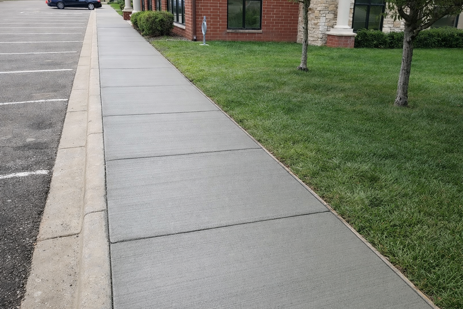 A concrete sidewalk with a grassy area on one side and a parking lot on the other.