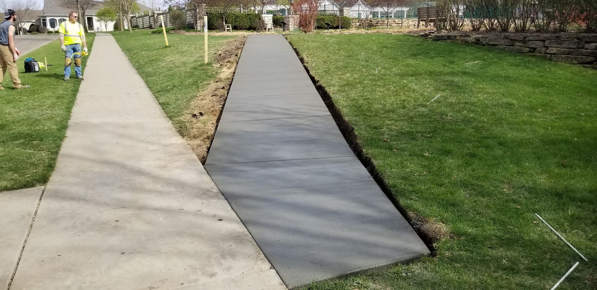 Newly poured concrete pathway next to an existing sidewalk; a person with tools in the background.