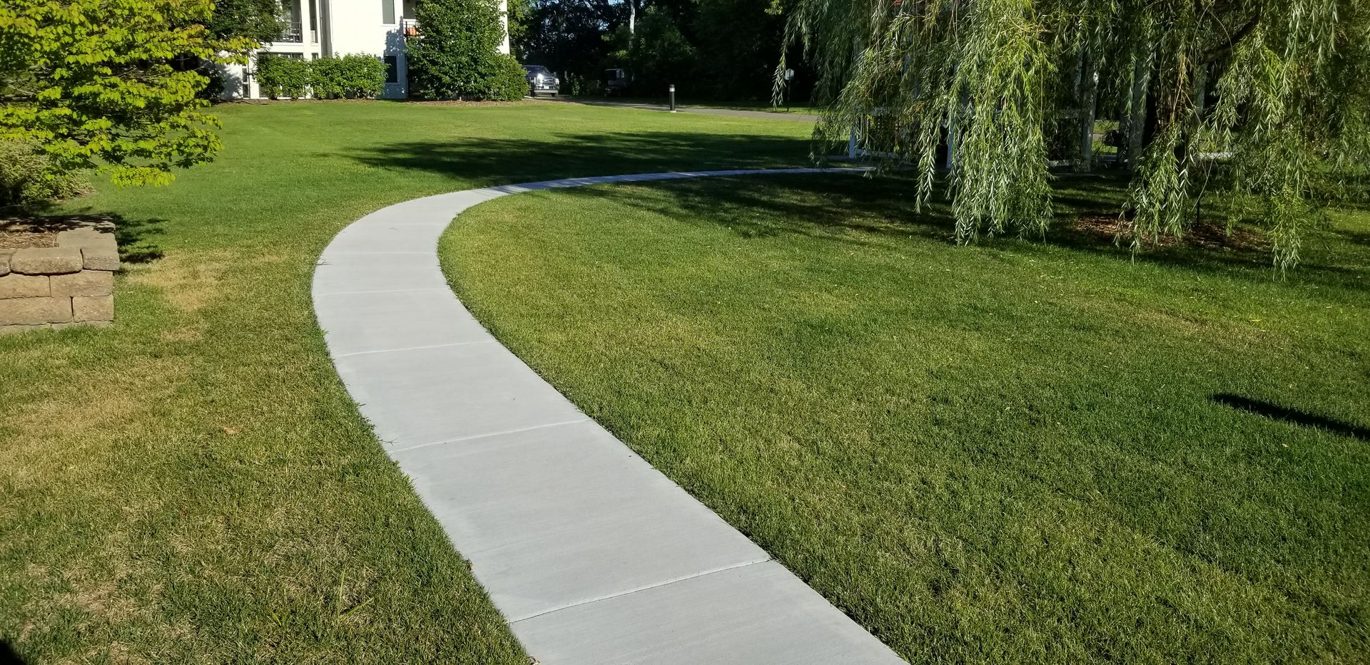Curving concrete path through green lawn, leading towards a white house and weeping willow tree.
