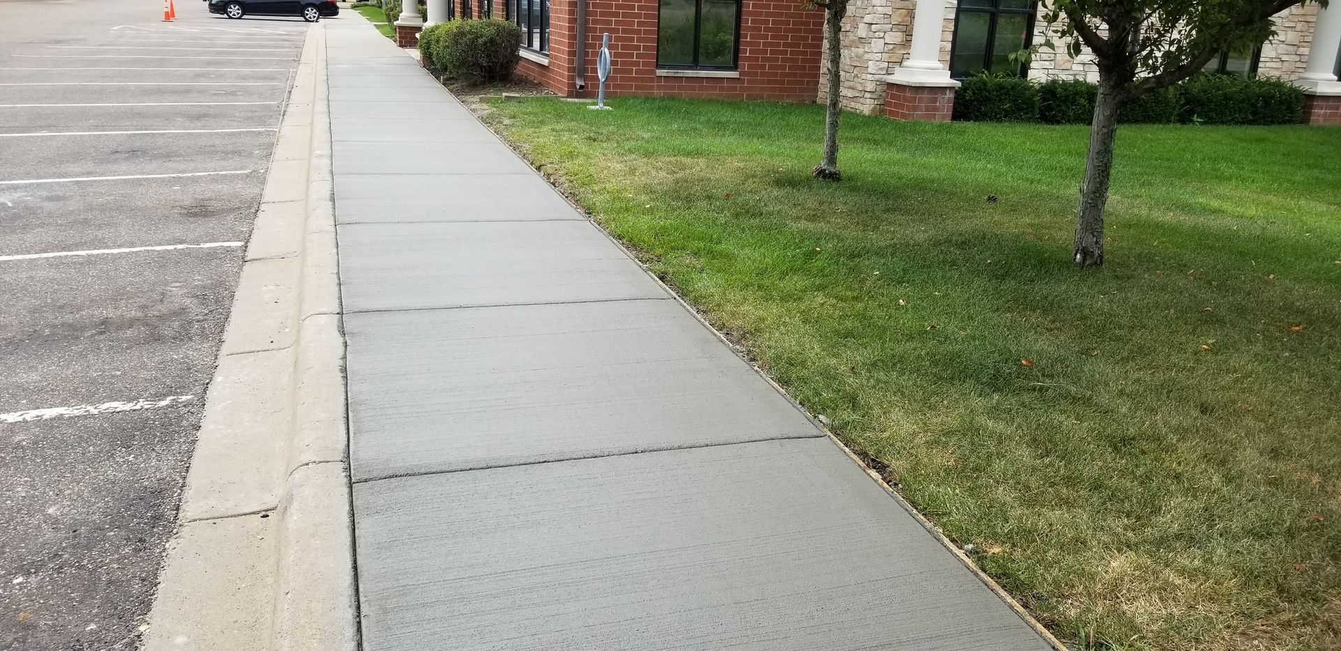 Sidewalk next to a parking lot and a grassy area with a building in the background.