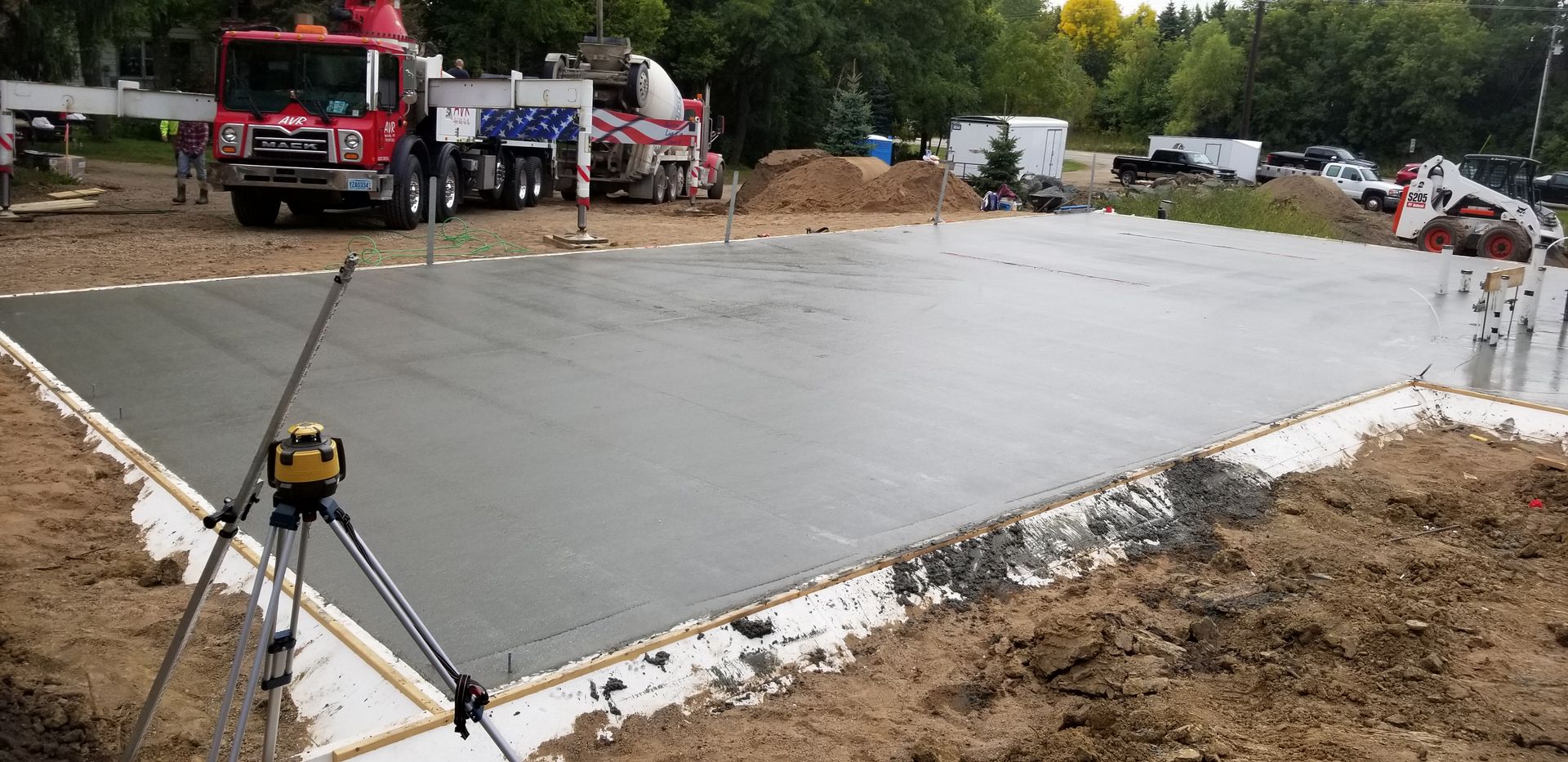 Freshly poured concrete foundation with cement trucks in the background and a leveling instrument in the foreground.