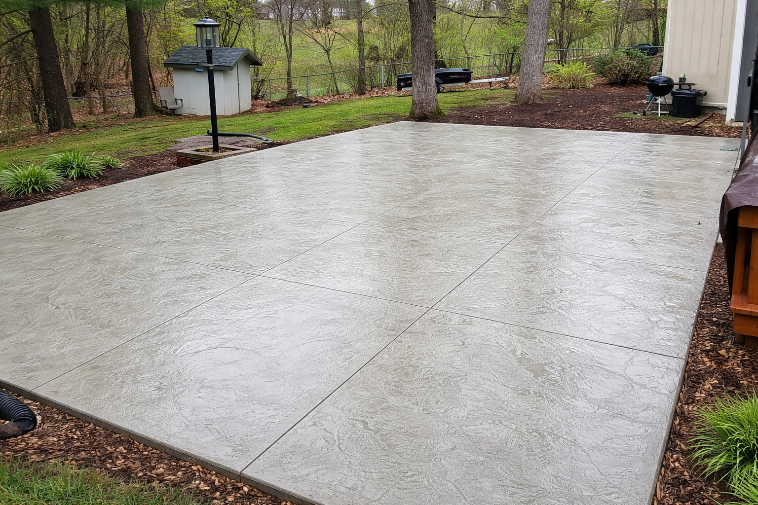 A gray concrete patio in a backyard setting, surrounded by grass and mulch beds.