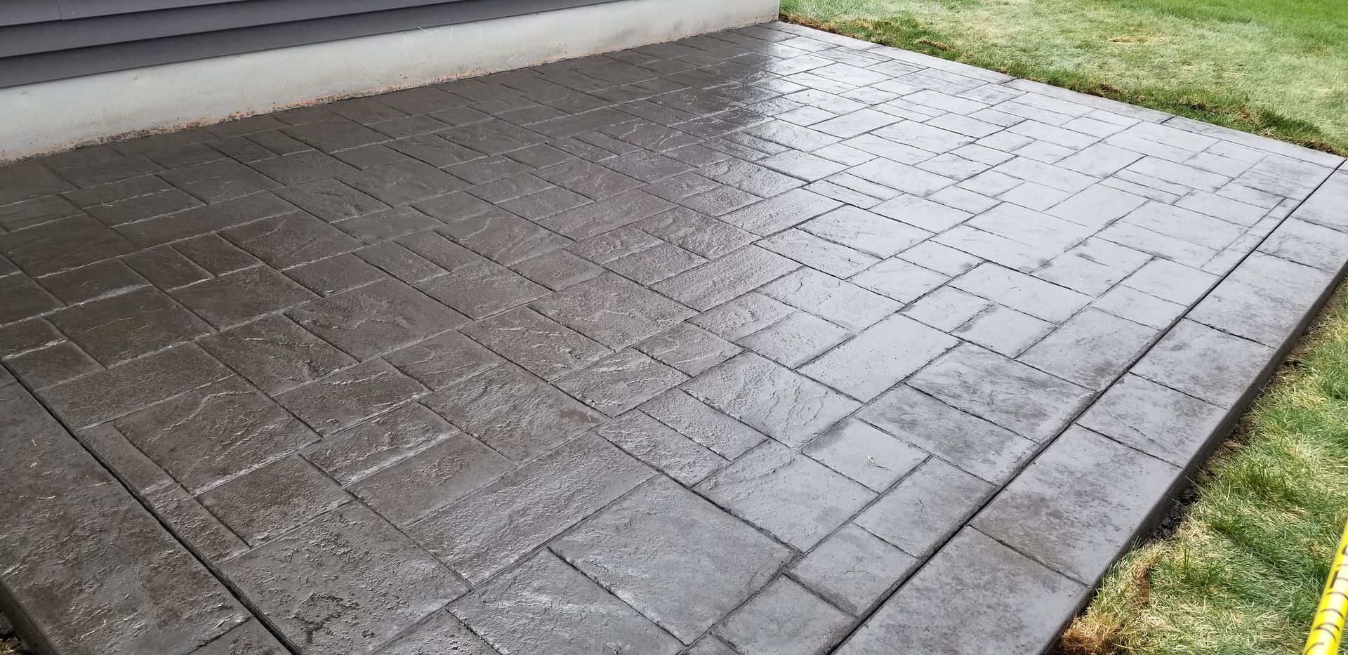 Stamped concrete patio with a brick pattern, wet from rain, next to green grass.