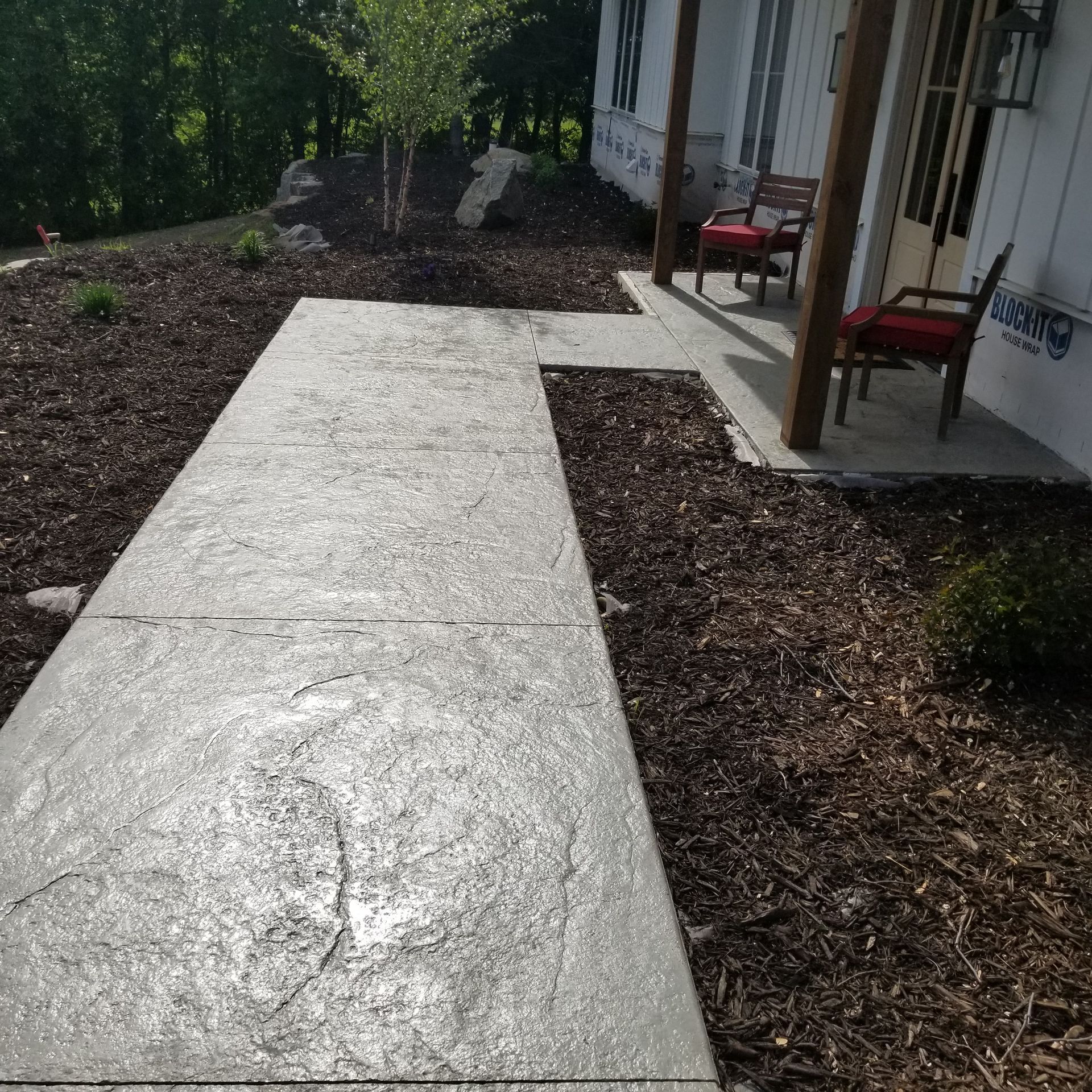 Concrete walkway leading to a porch with two chairs, surrounded by brown mulch and landscaping.