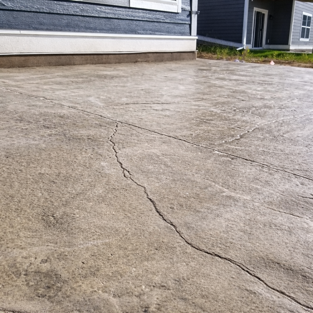 Cracked concrete driveway with a light brown color. The crack is dark and jagged. Adjacent is a blue house.