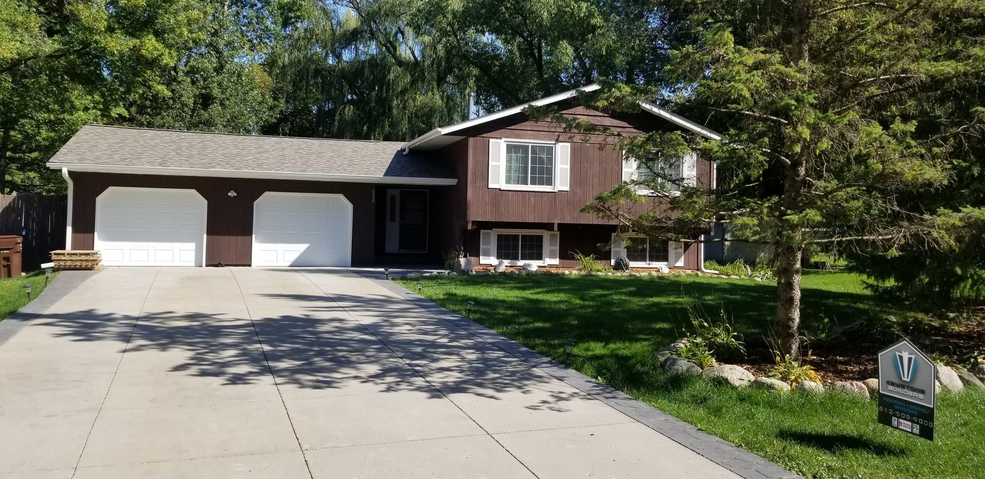 Brown house with white garage doors and a paved driveway. Trees and green grass surround it.