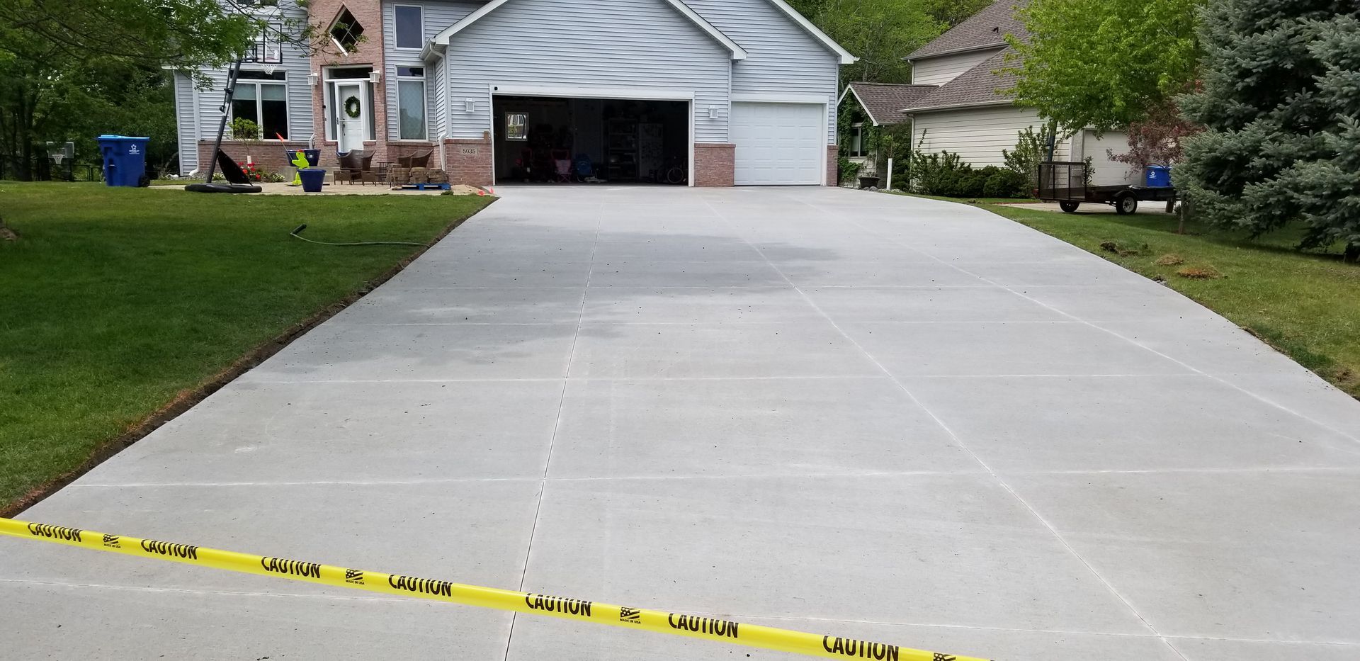 A long gray concrete driveway leads to a house with a garage. Yellow caution tape is across the bottom.