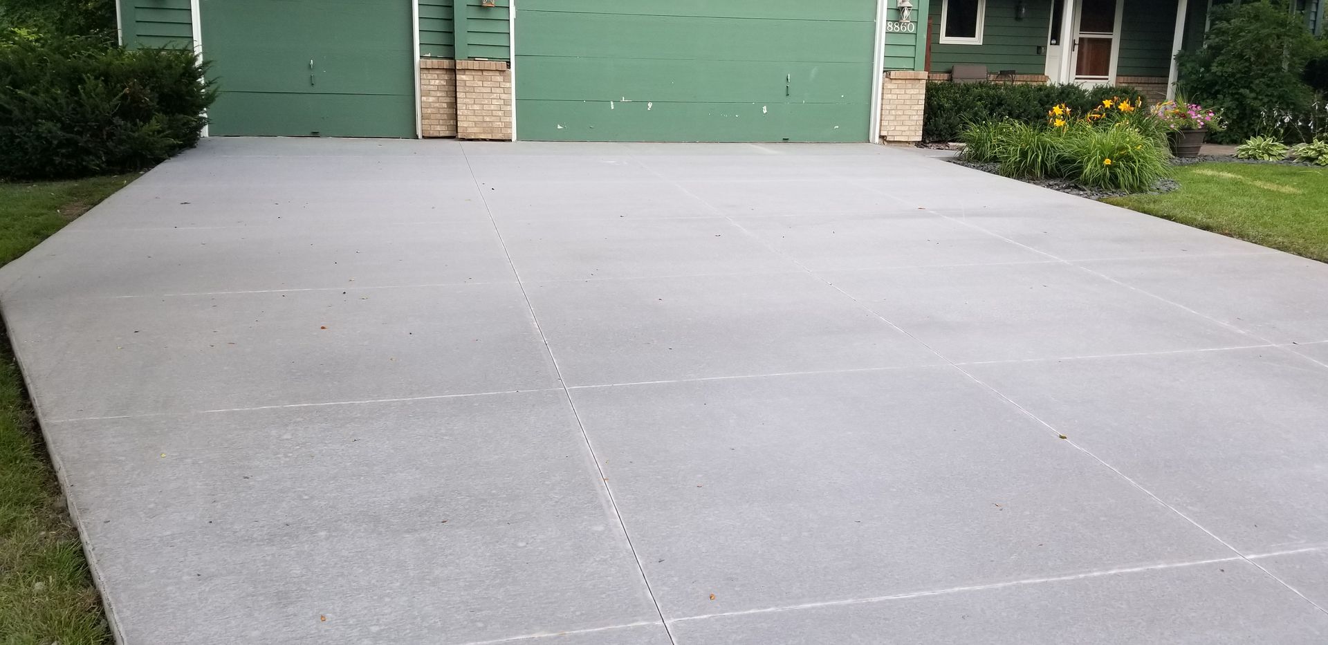 A gray concrete driveway in front of a green garage, with a house visible in the background.