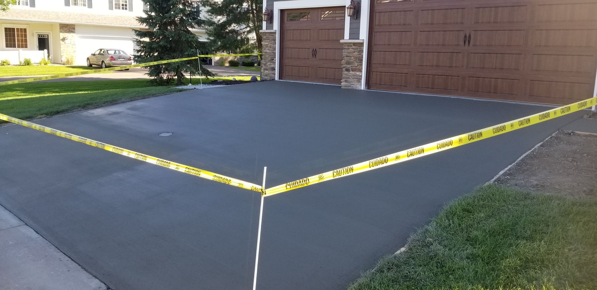 Freshly paved asphalt driveway with yellow caution tape, next to grass lawn and a house.