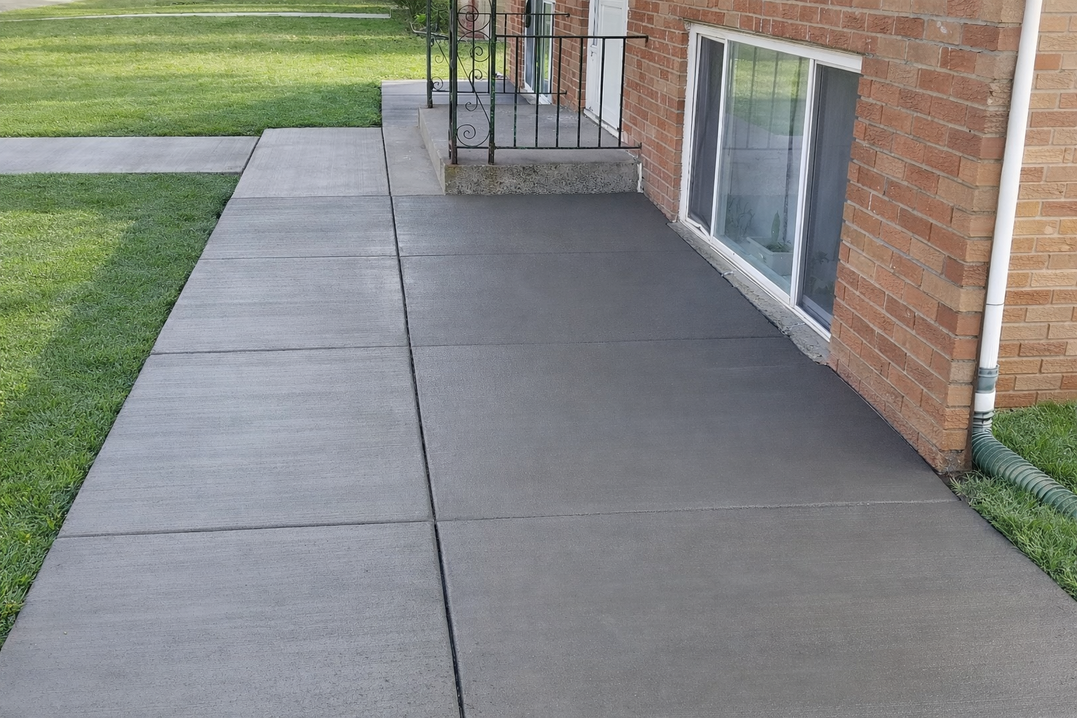 Concrete sidewalk and porch next to a brick building and grass lawn.