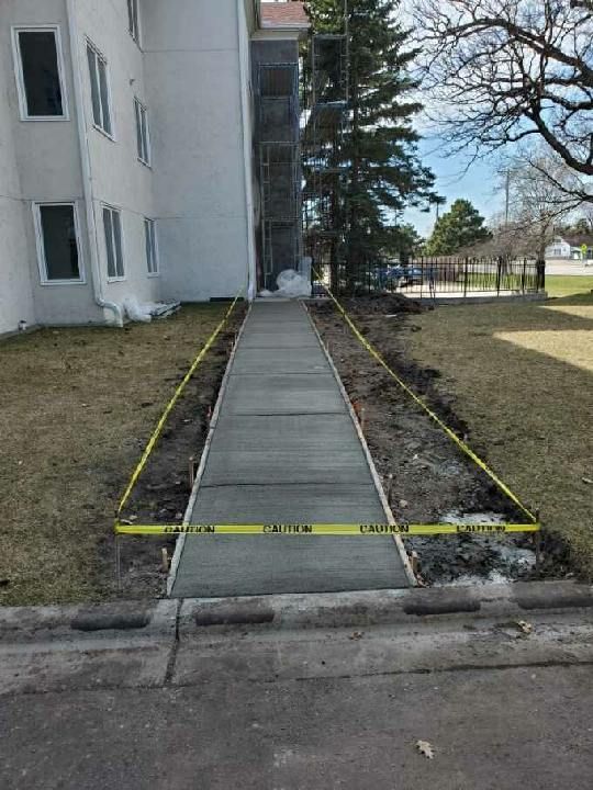 New concrete sidewalk with yellow caution tape, adjacent to building and grass.