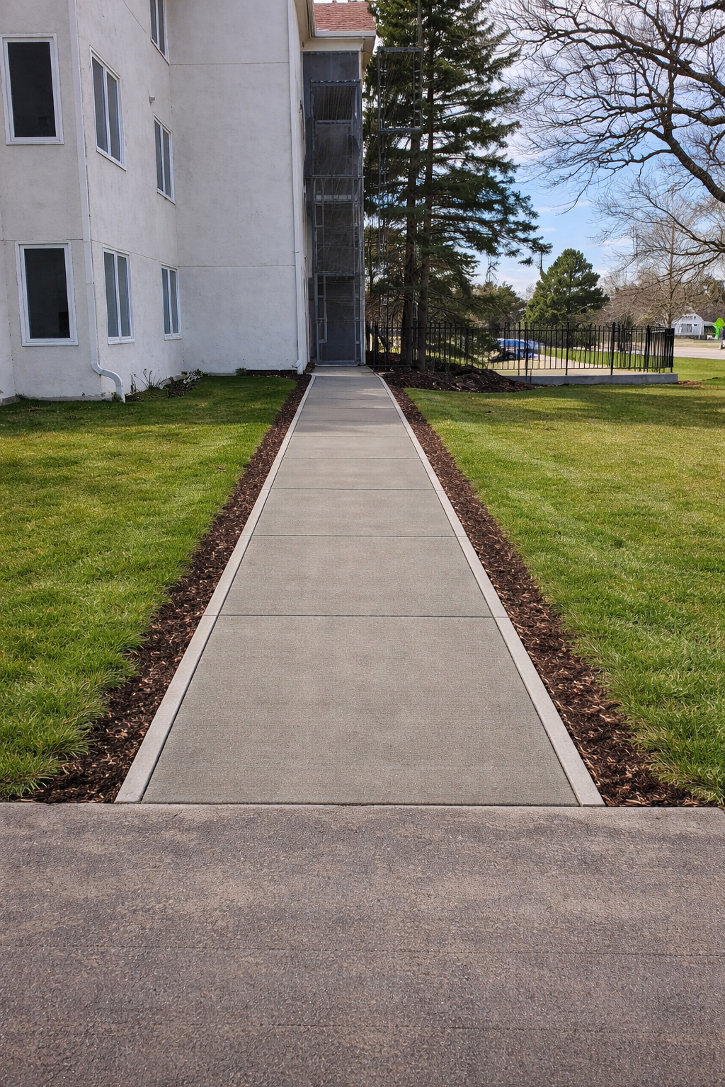 A concrete pathway leads to a building entrance, bordered by grass and mulch. Sunny day.