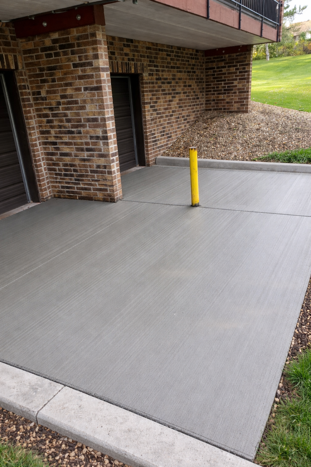 Gray concrete parking area next to a brick building and grass. A yellow bollard stands in the middle.