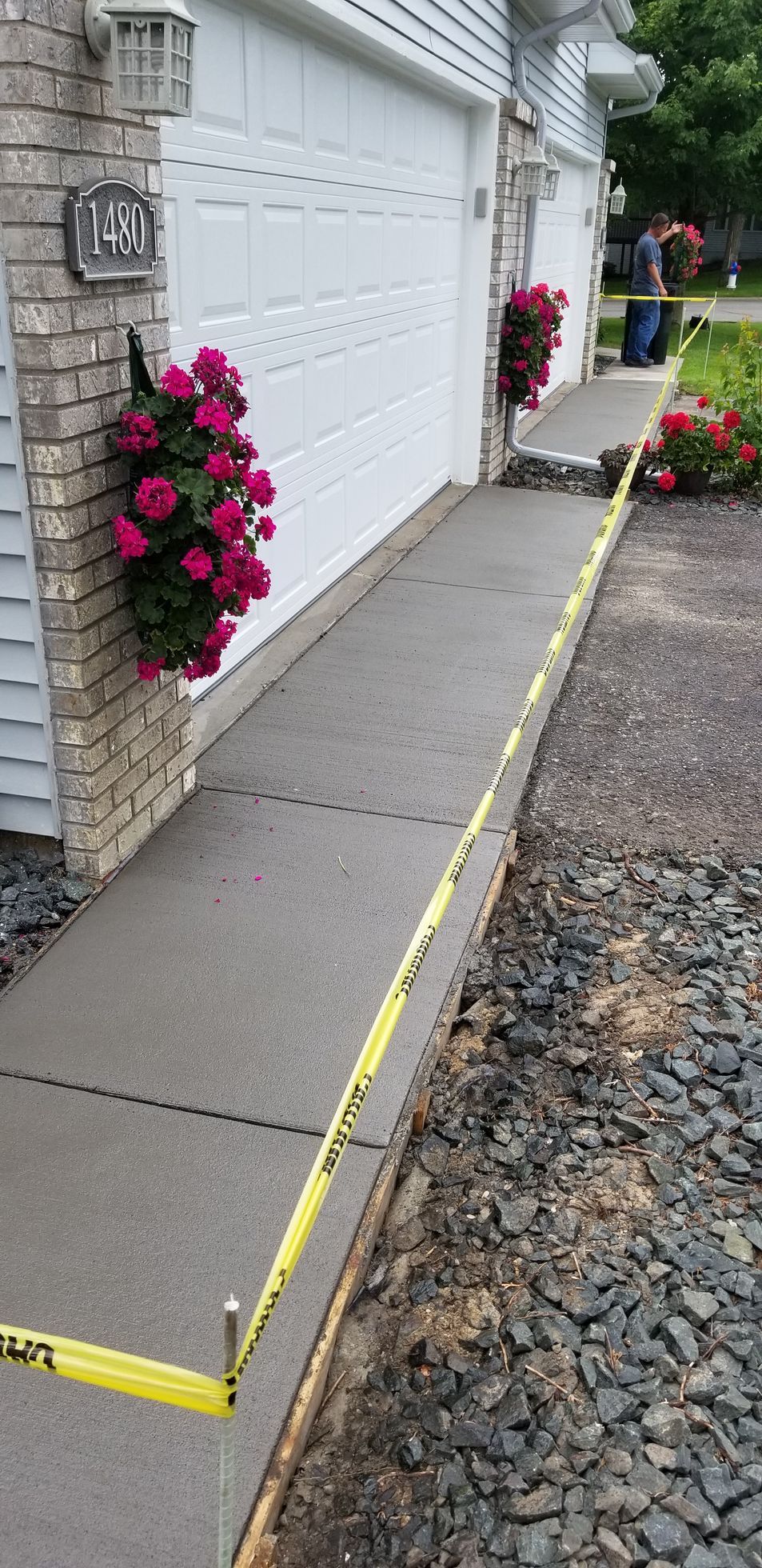 New concrete sidewalk with caution tape, next to gravel driveway, house exterior in the background.