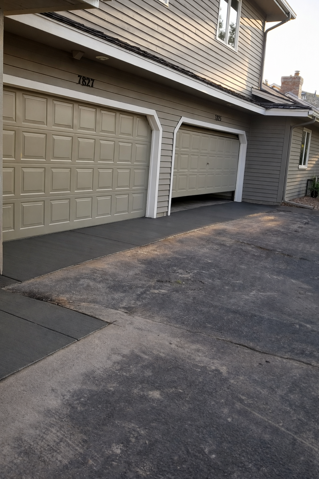 Two-car garage with one door partially open. Tan garage doors, gray siding, asphalt driveway.
