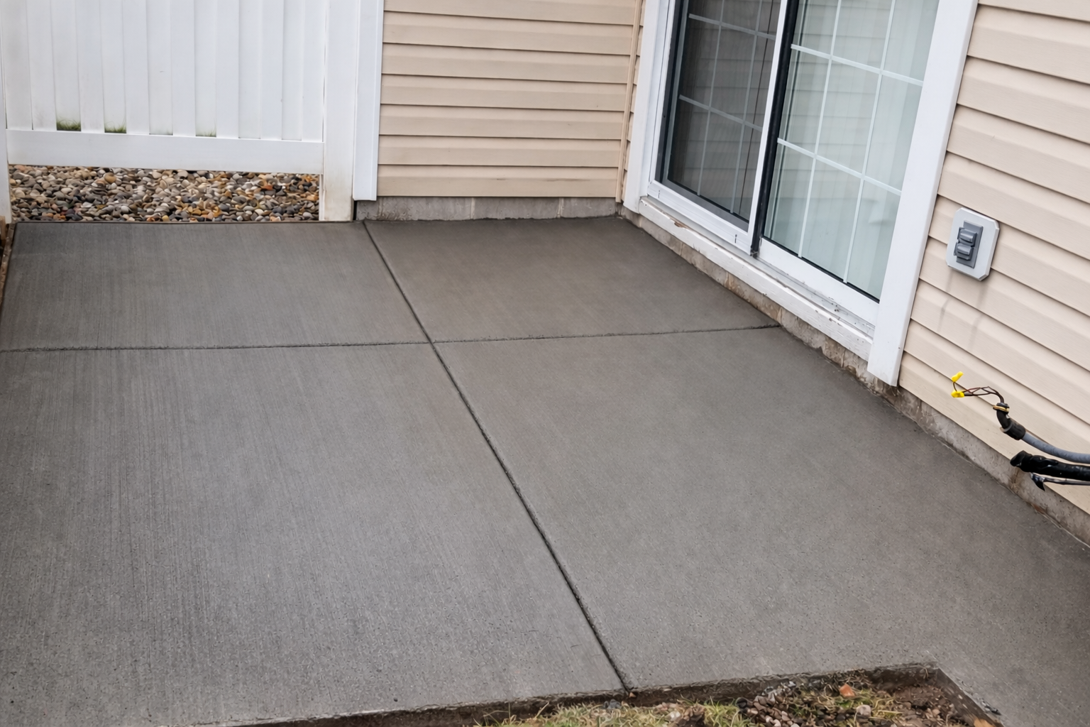 A gray concrete patio outside a building with a sliding glass door and white fence.