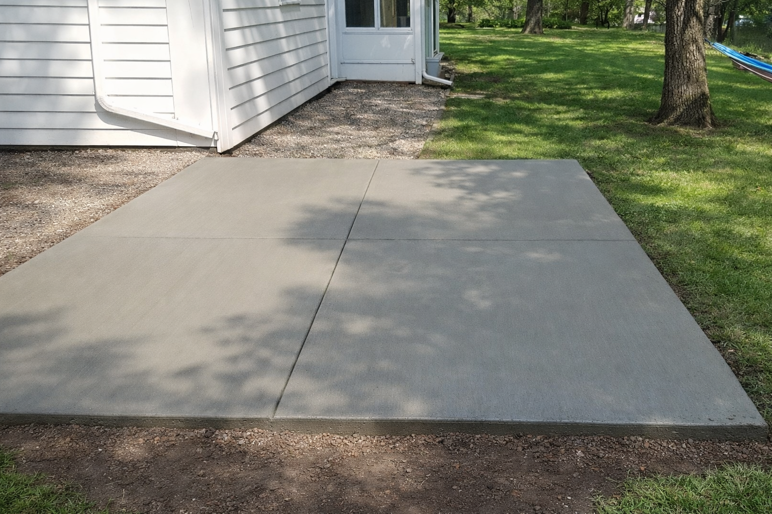 Concrete patio next to a white house with a grassy yard and a tree.