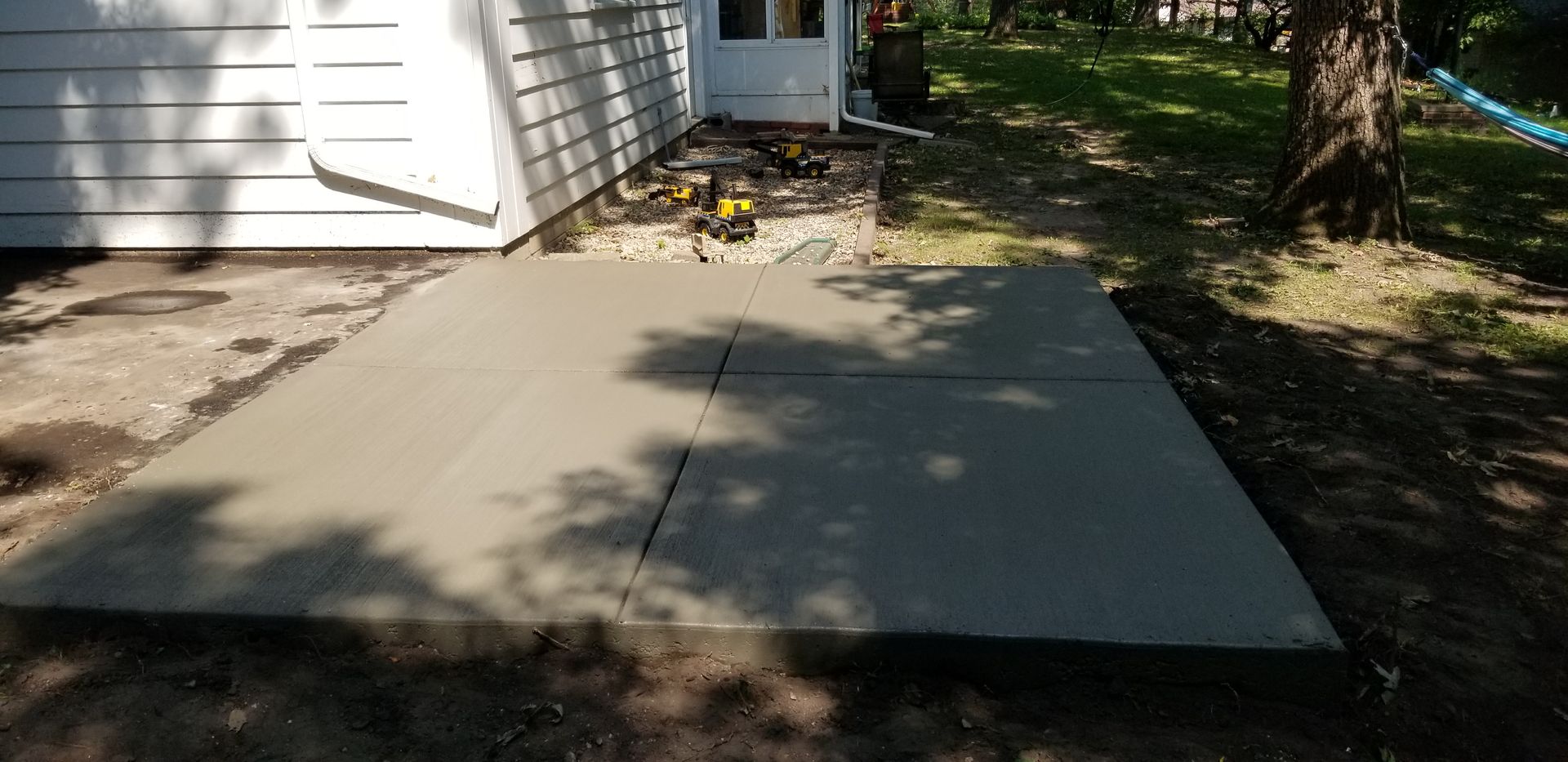 Newly poured concrete patio next to a white house with a surrounding grassy area and a tree.