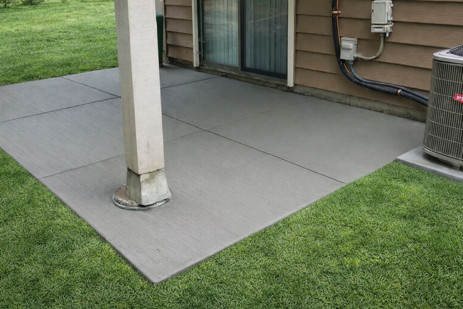 Concrete patio with a support column and air conditioning unit, next to a building and lawn.