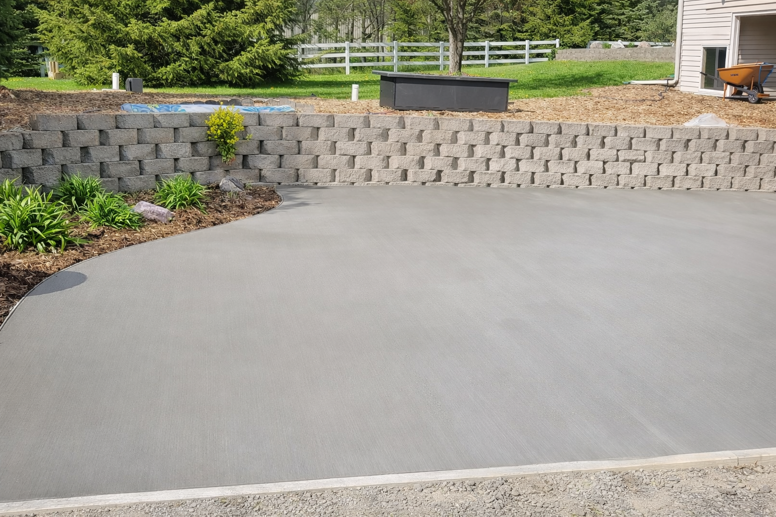 Newly poured concrete patio in front of a retaining wall made of textured gray blocks, and landscaping.