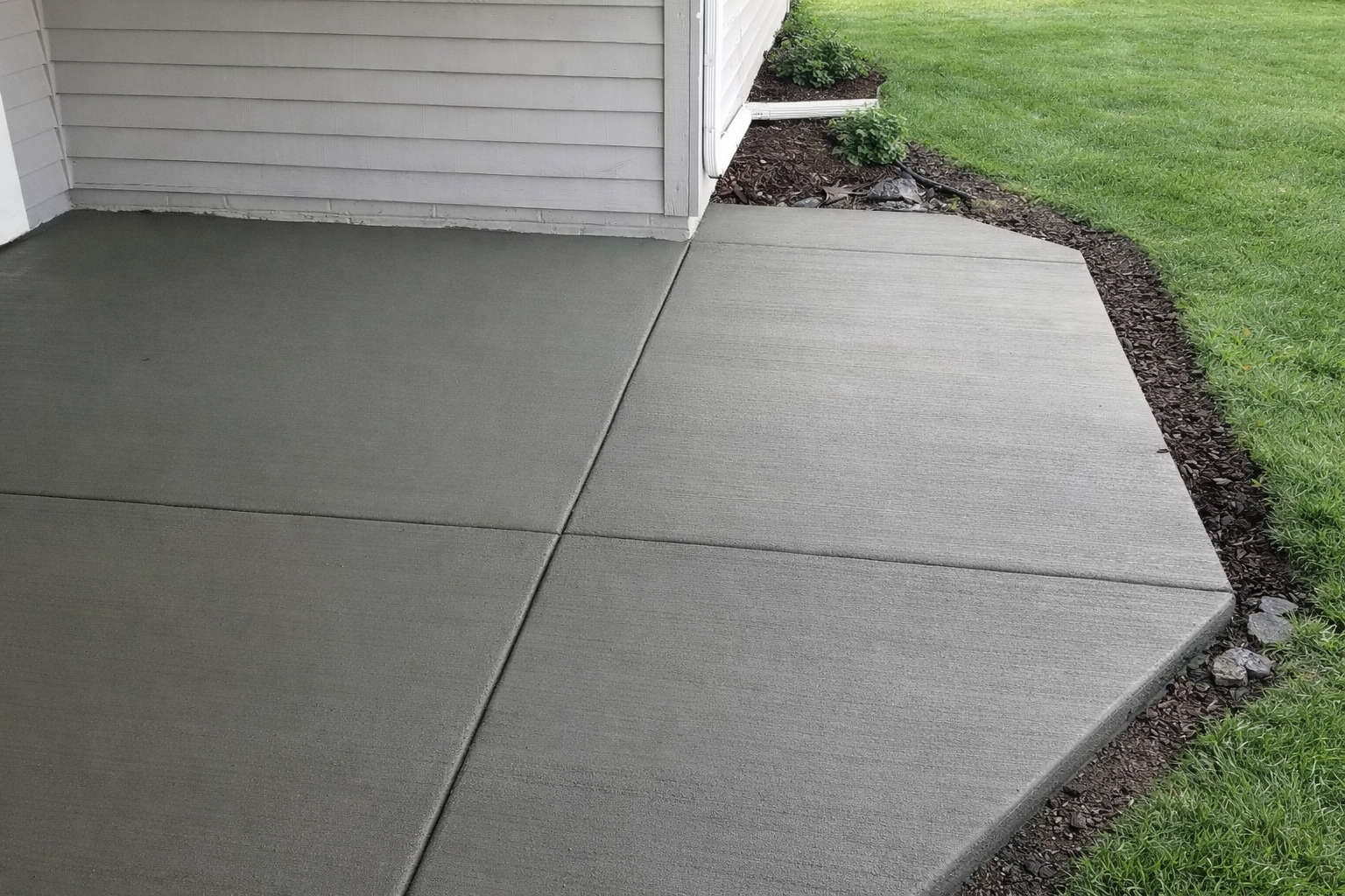 Gray concrete patio next to a grass lawn. Beige siding is above the patio.