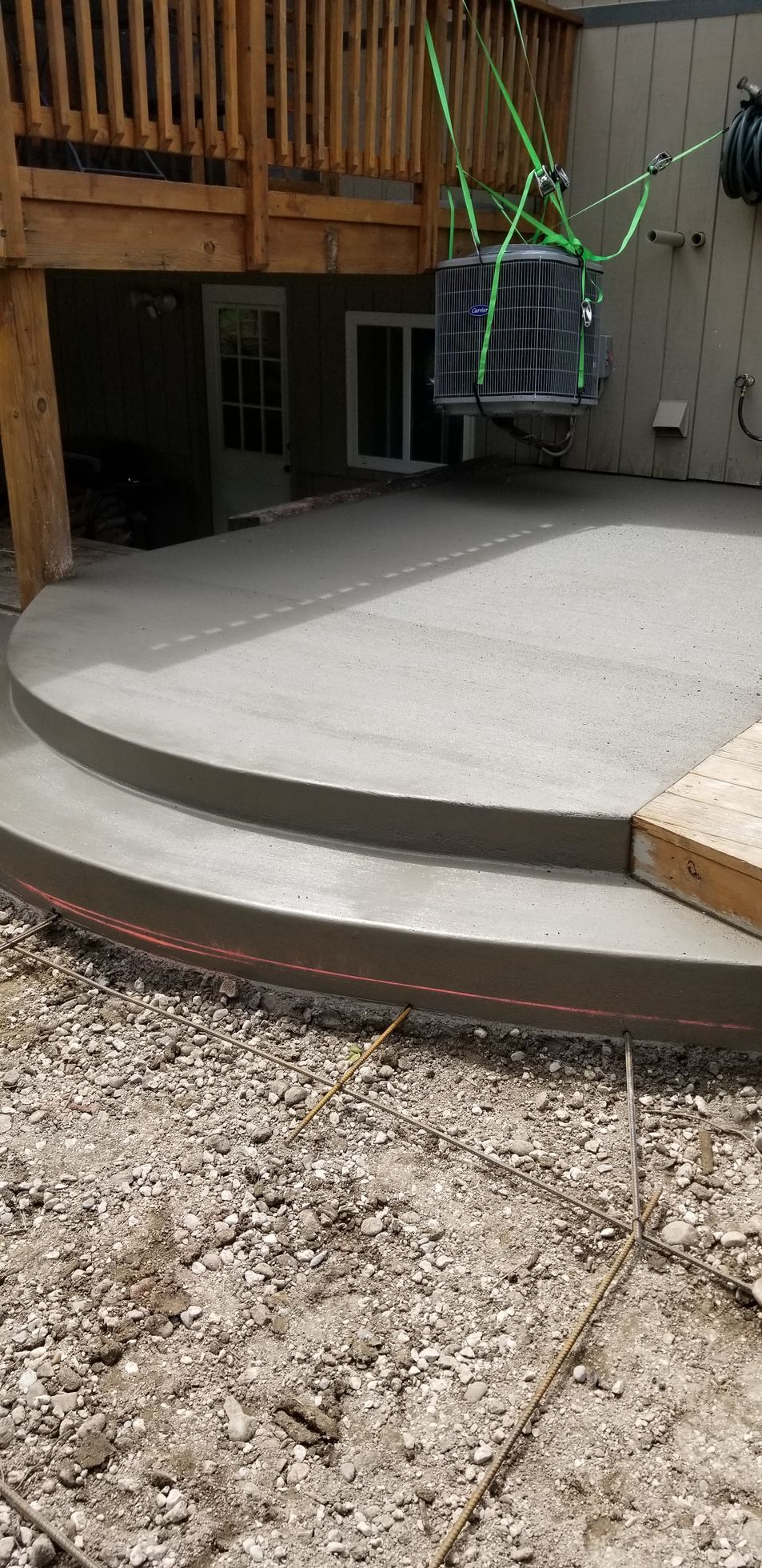 Concrete patio with steps being built, an AC unit suspended above by green straps.