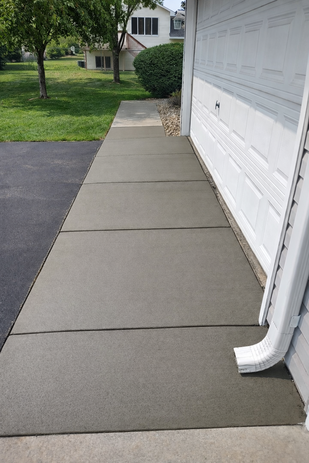 Newly poured concrete sidewalk next to a white garage, next to a driveway and lawn.