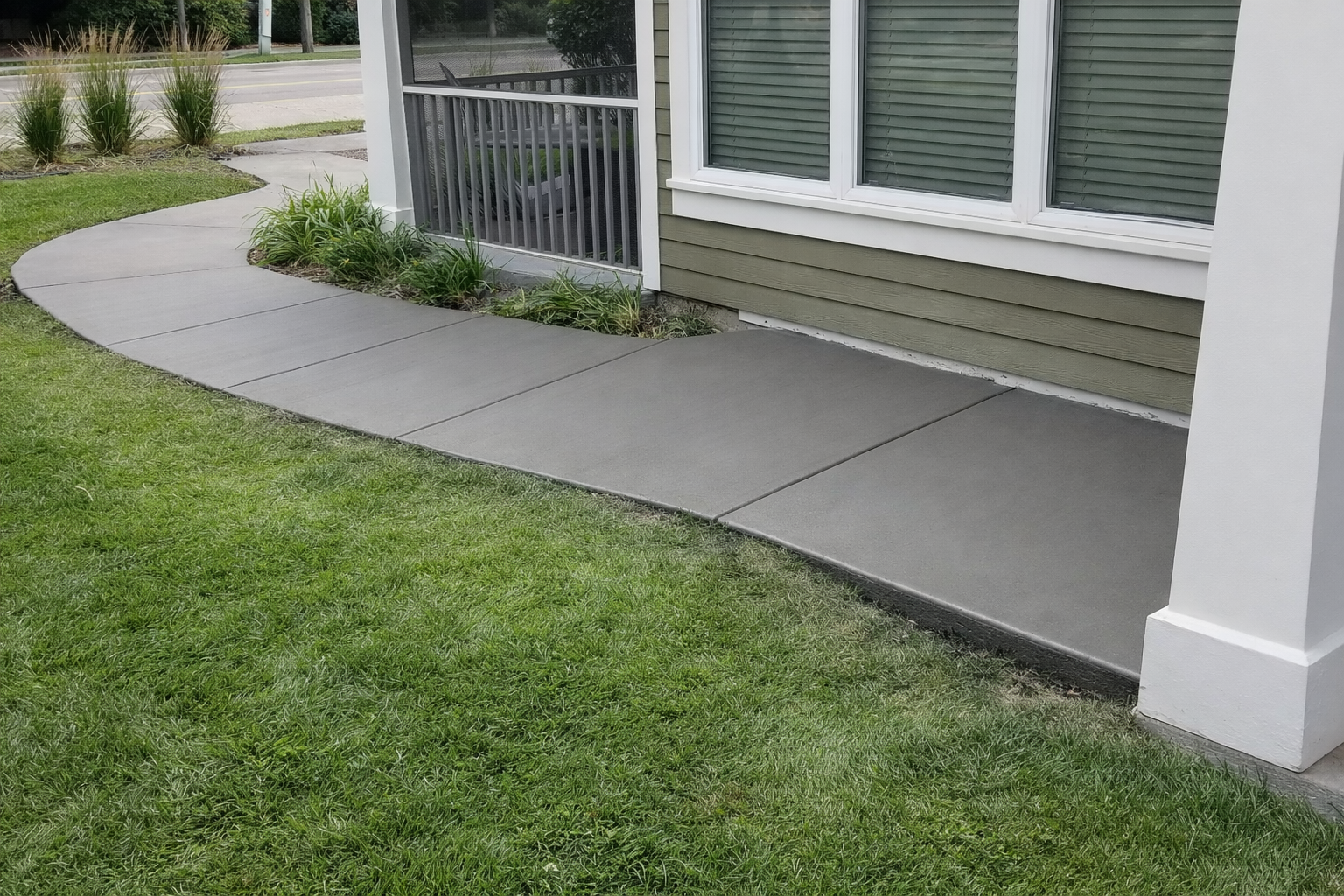 Gray concrete walkway curving along a house with green siding and grass.