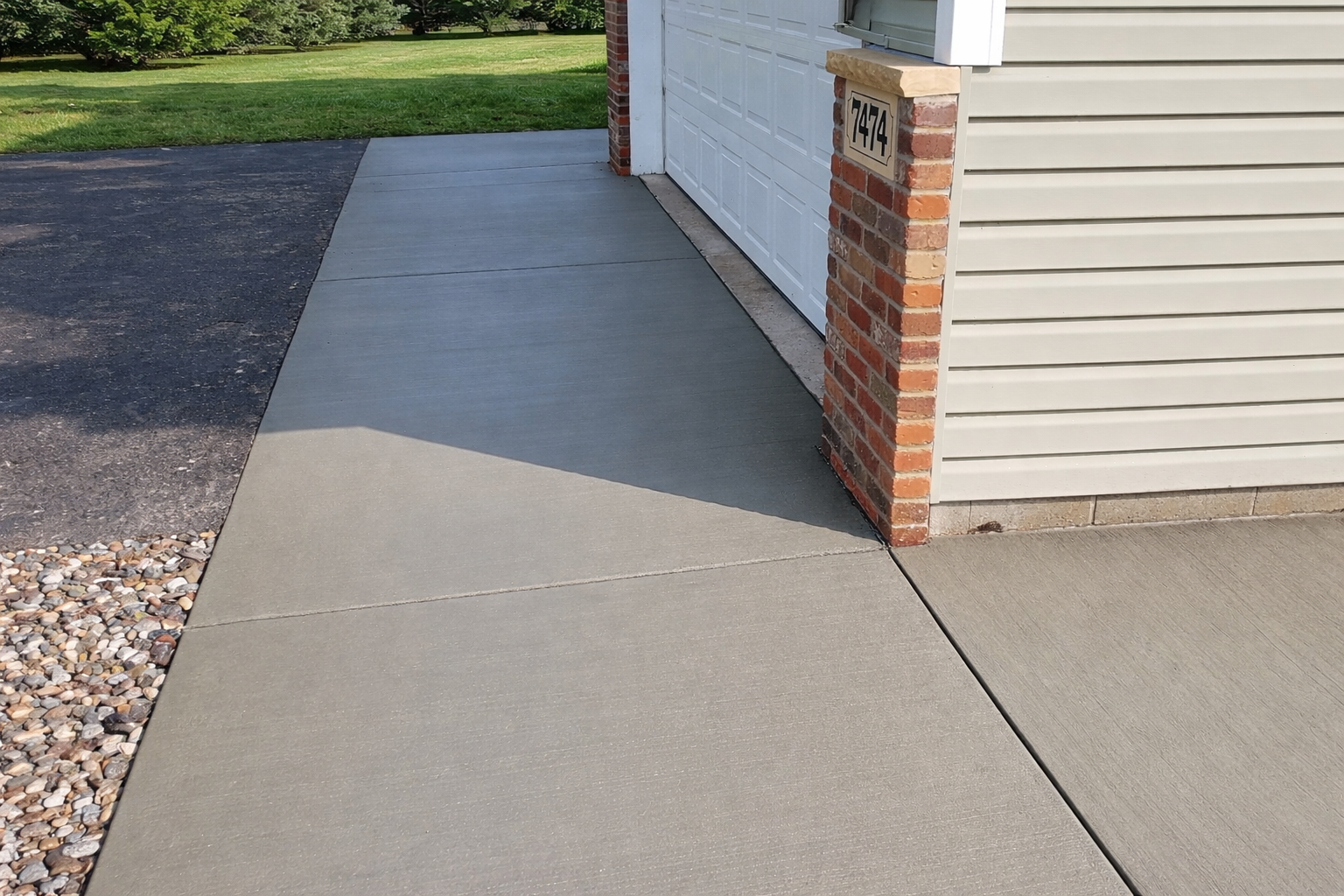 Freshly poured gray concrete sidewalk leading to a garage. Brick accent and beige siding visible.