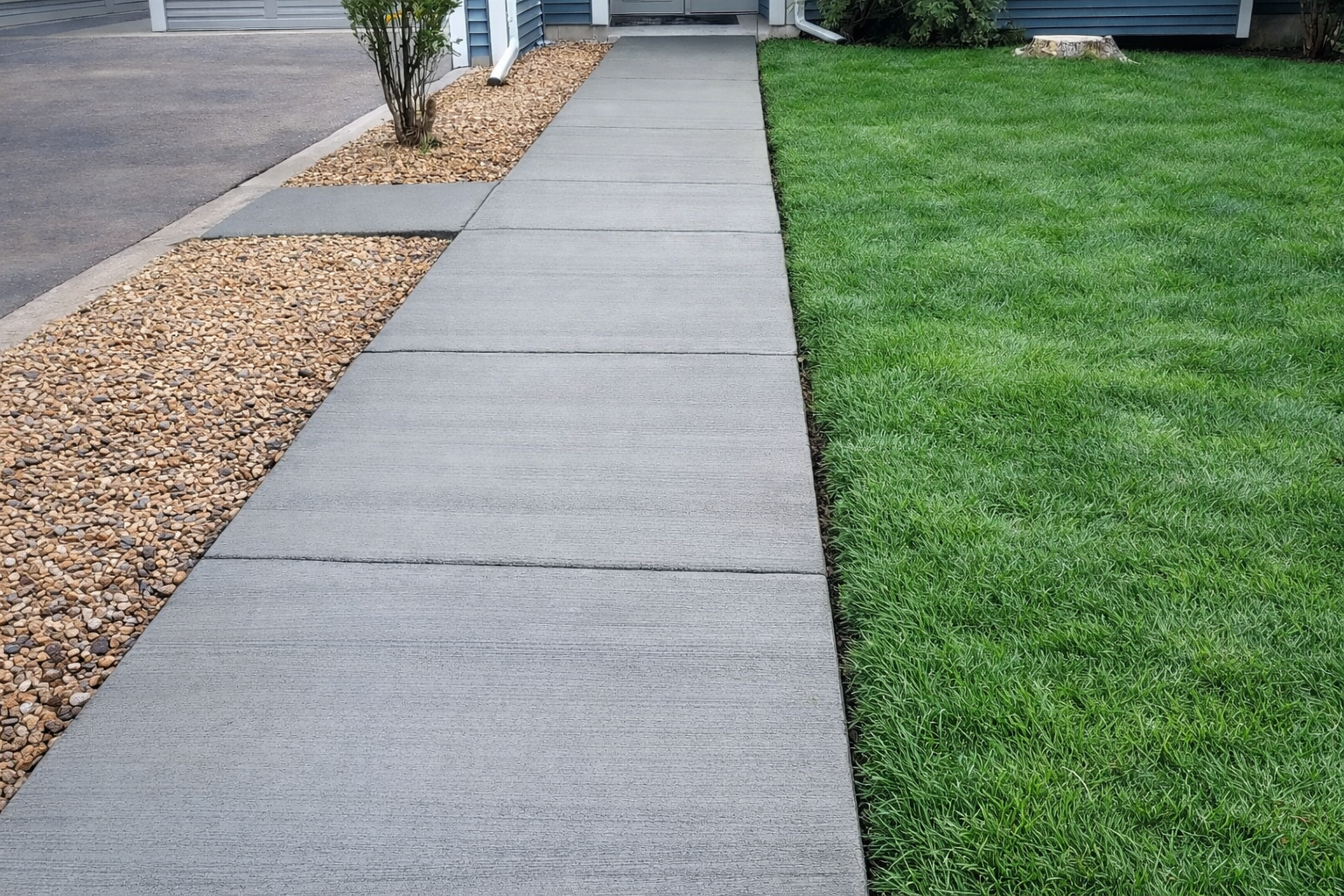 Concrete walkway bordered by brown gravel and green grass, leading to a house entrance.