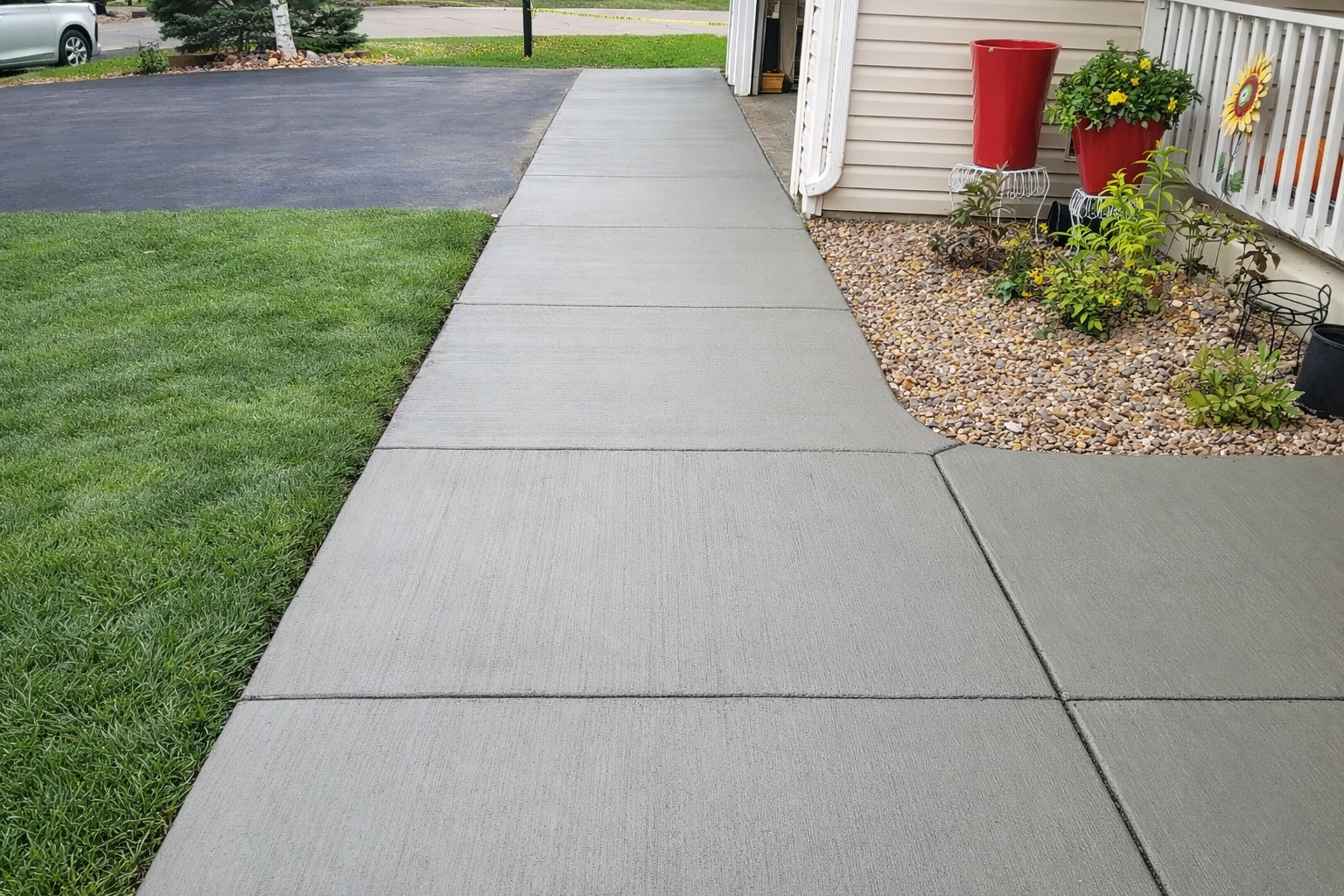 Newly poured concrete sidewalk next to a green lawn and a house with a small flower bed.