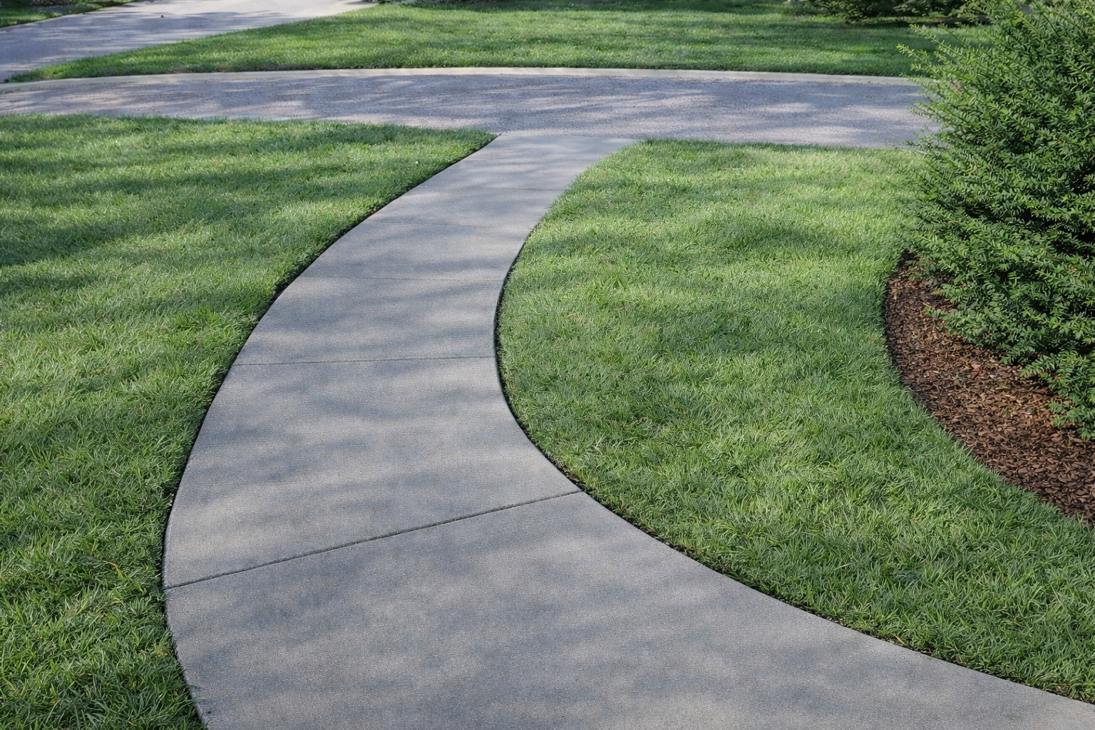 Curving concrete pathway through a grassy yard, next to a bush and another paved area.