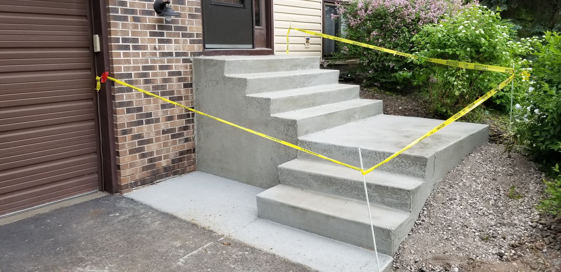 Concrete steps with yellow caution tape are blocked off near a garage and bushes.