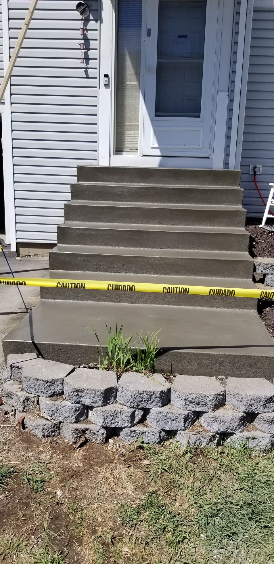 Concrete steps leading to a white door. A yellow caution tape is across the bottom steps. A retaining wall and grass are in the foreground.