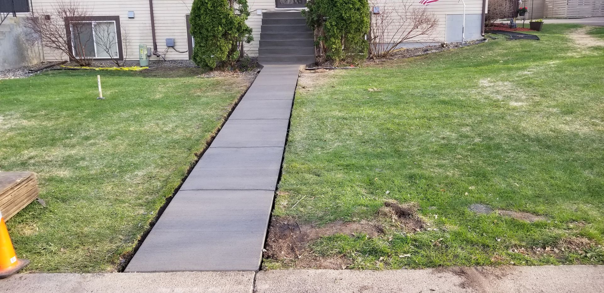 A concrete walkway leads to a house with steps, flanked by green grass and two evergreen trees.
