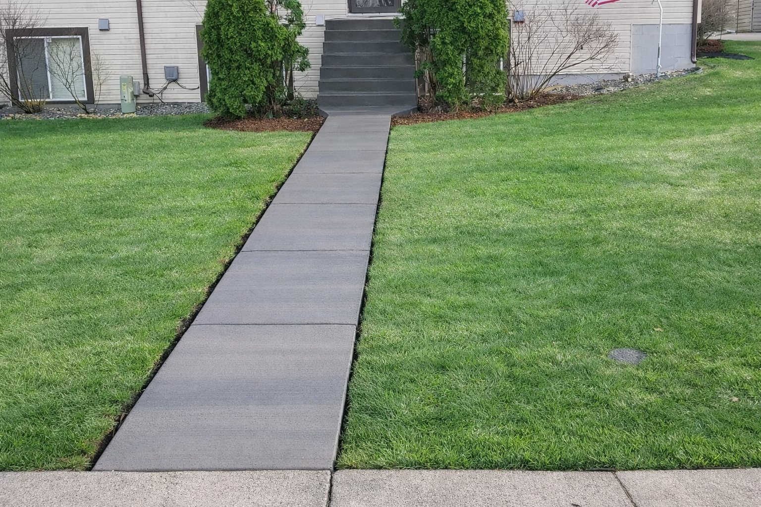 Gray concrete walkway leading to front steps of a house, surrounded by green grass.