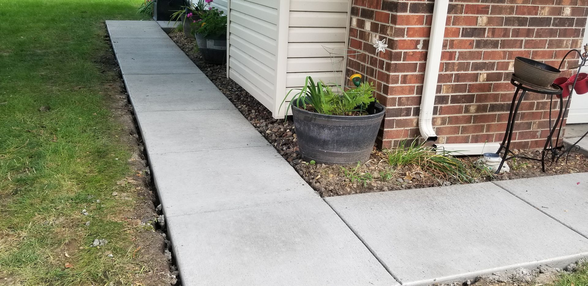 Concrete walkway alongside a building and grass.  Dark planter with plants at the building's edge.