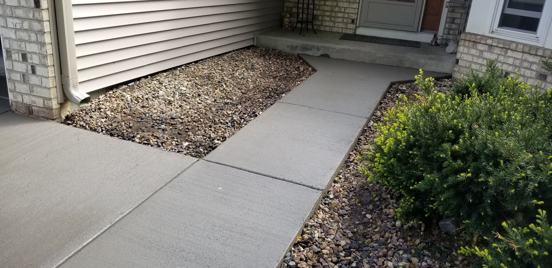 Concrete walkway leading to a front door, next to a rock bed and green bush.