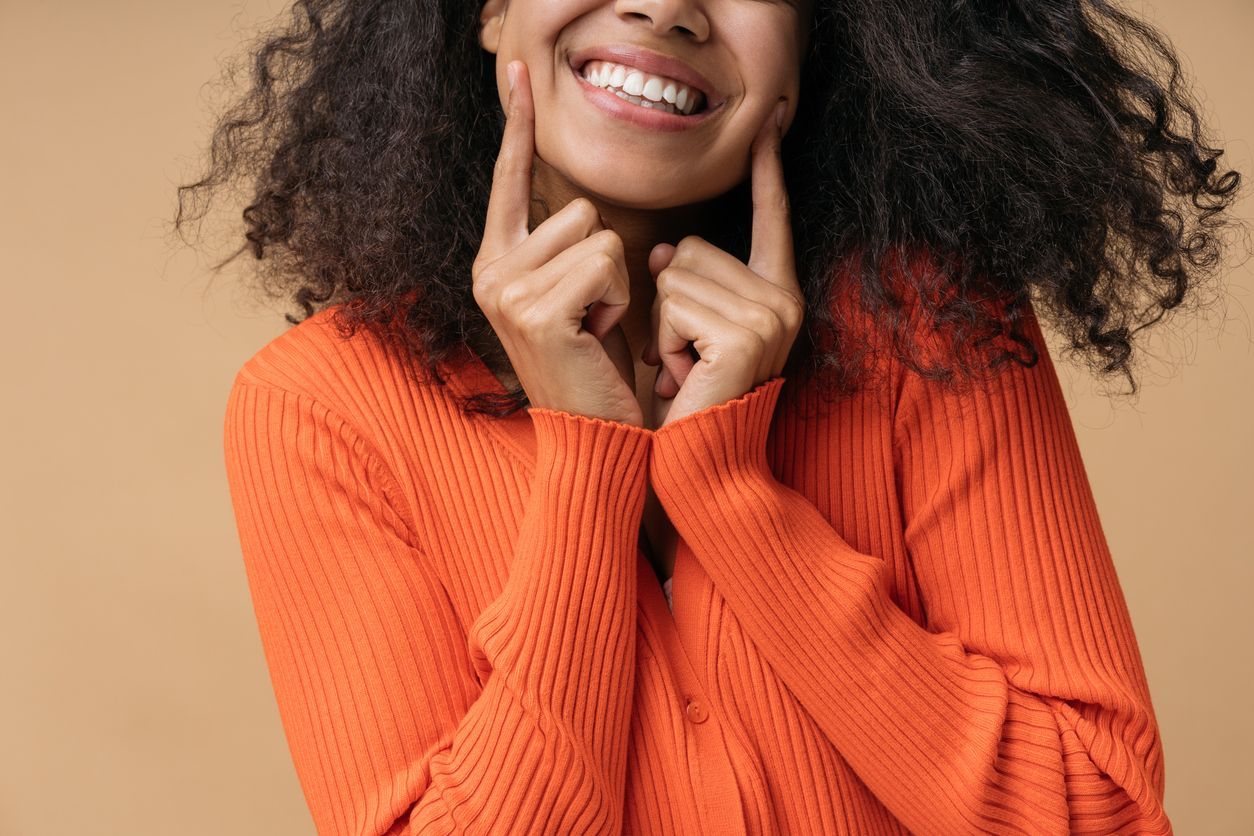 A woman in an orange sweater is smiling and touching her face.