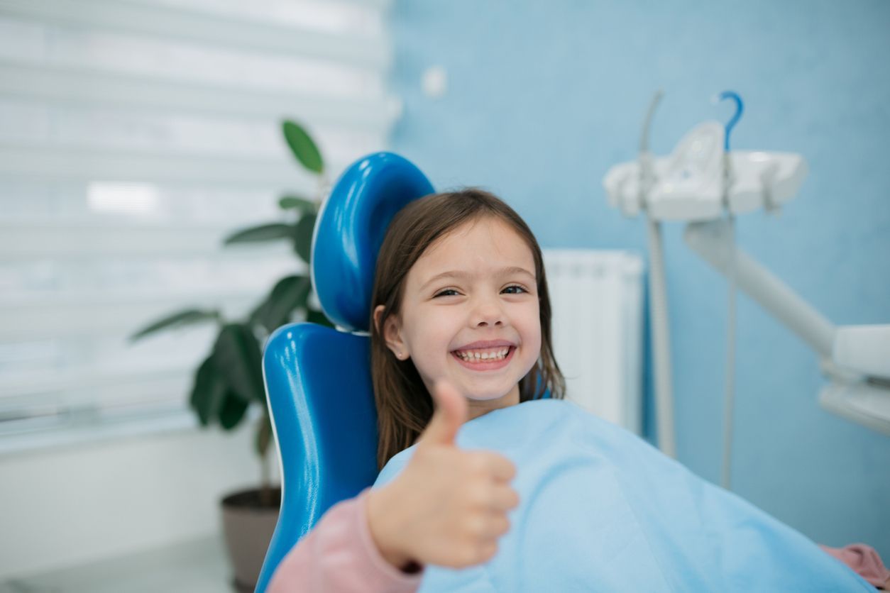 A little girl is sitting in a dental chair and giving a thumbs up.