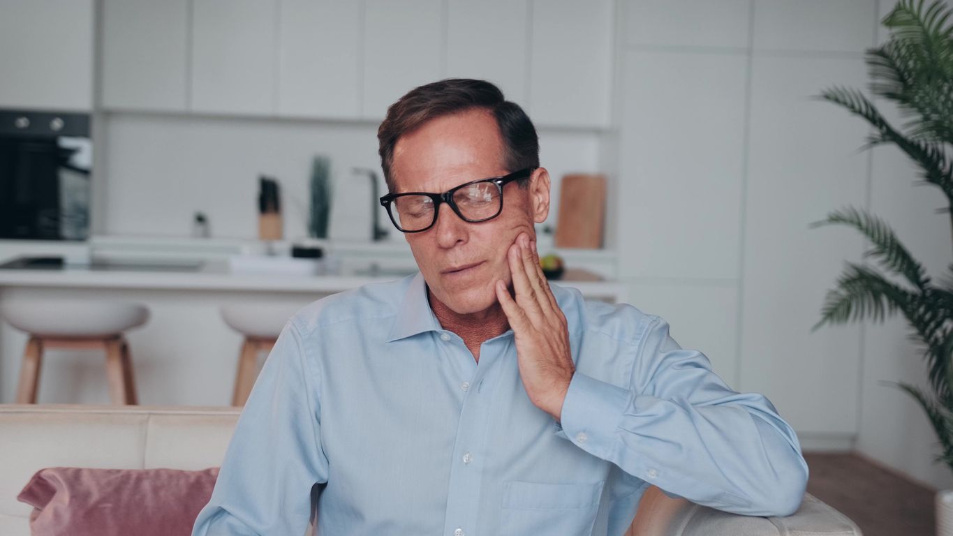 A man with glasses is sitting on a couch holding his face in pain.