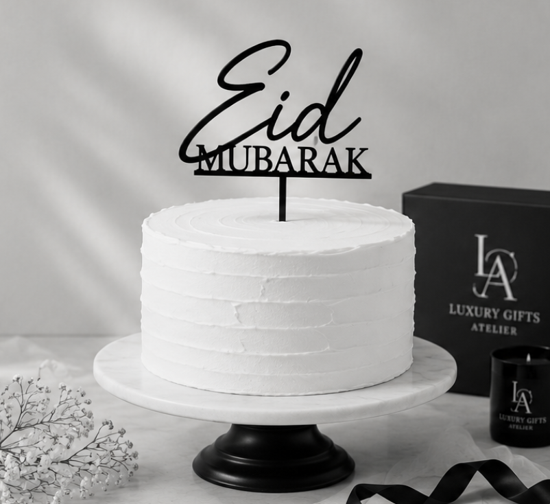 Witte taart met Eid Mubarak-decoratie op een standaard, naast luxe zwarte geschenkdozen op een grijze achtergrond.