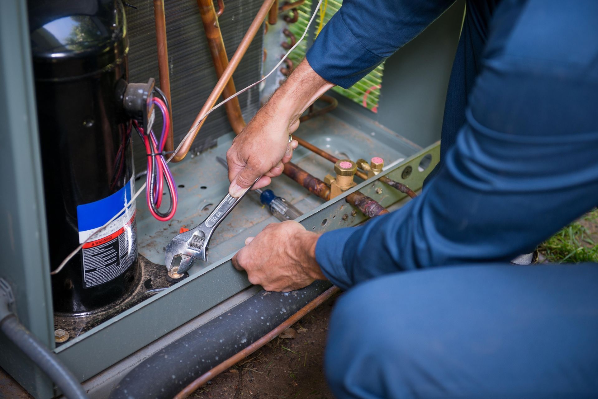 Person in blue suit repairs outdoor AC unit with a wrench.